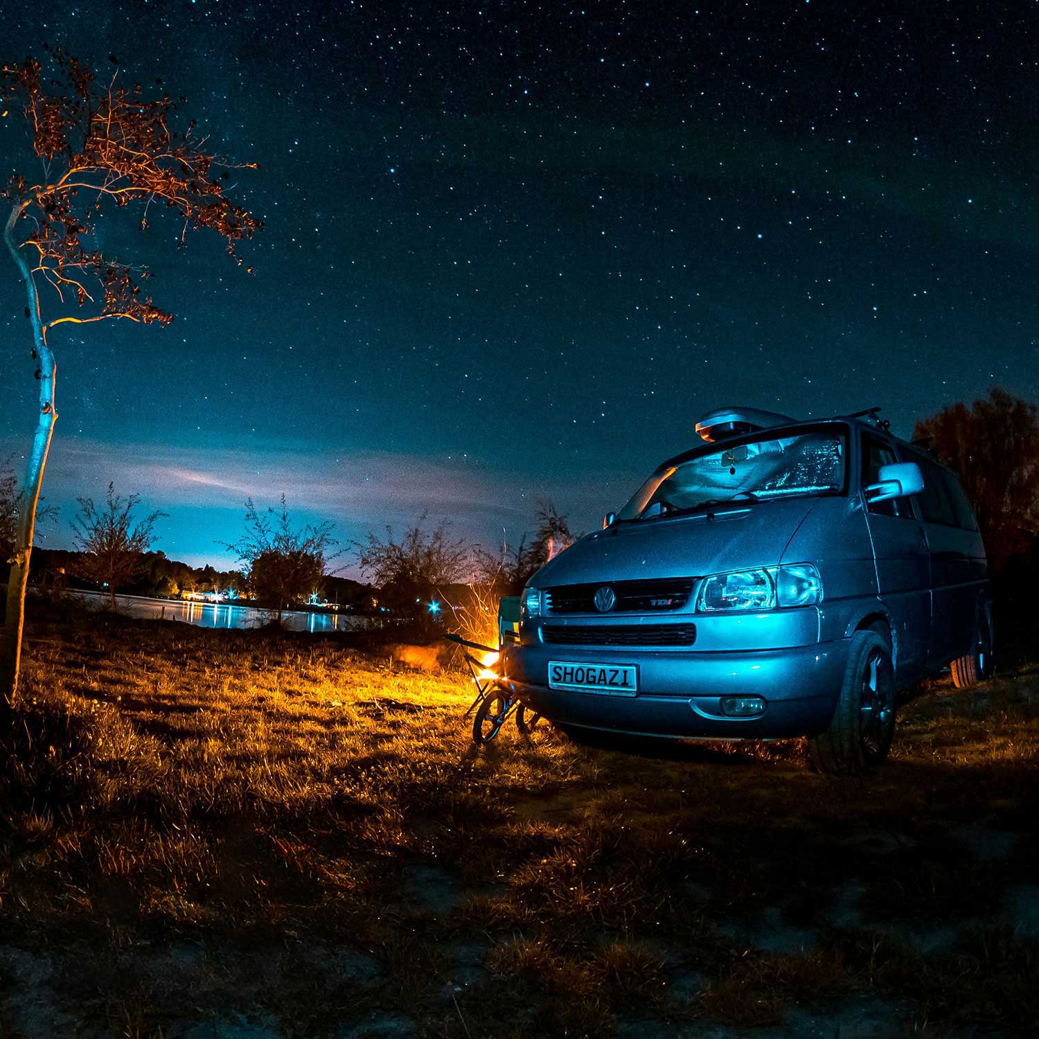 VW T4 steht nachts auf einer Wiese am See unter klarem Sternenhimmel, mit einer Lichtquelle oder Feuerstelle im Vordergrund – Matratze 120x186 cm für Camping