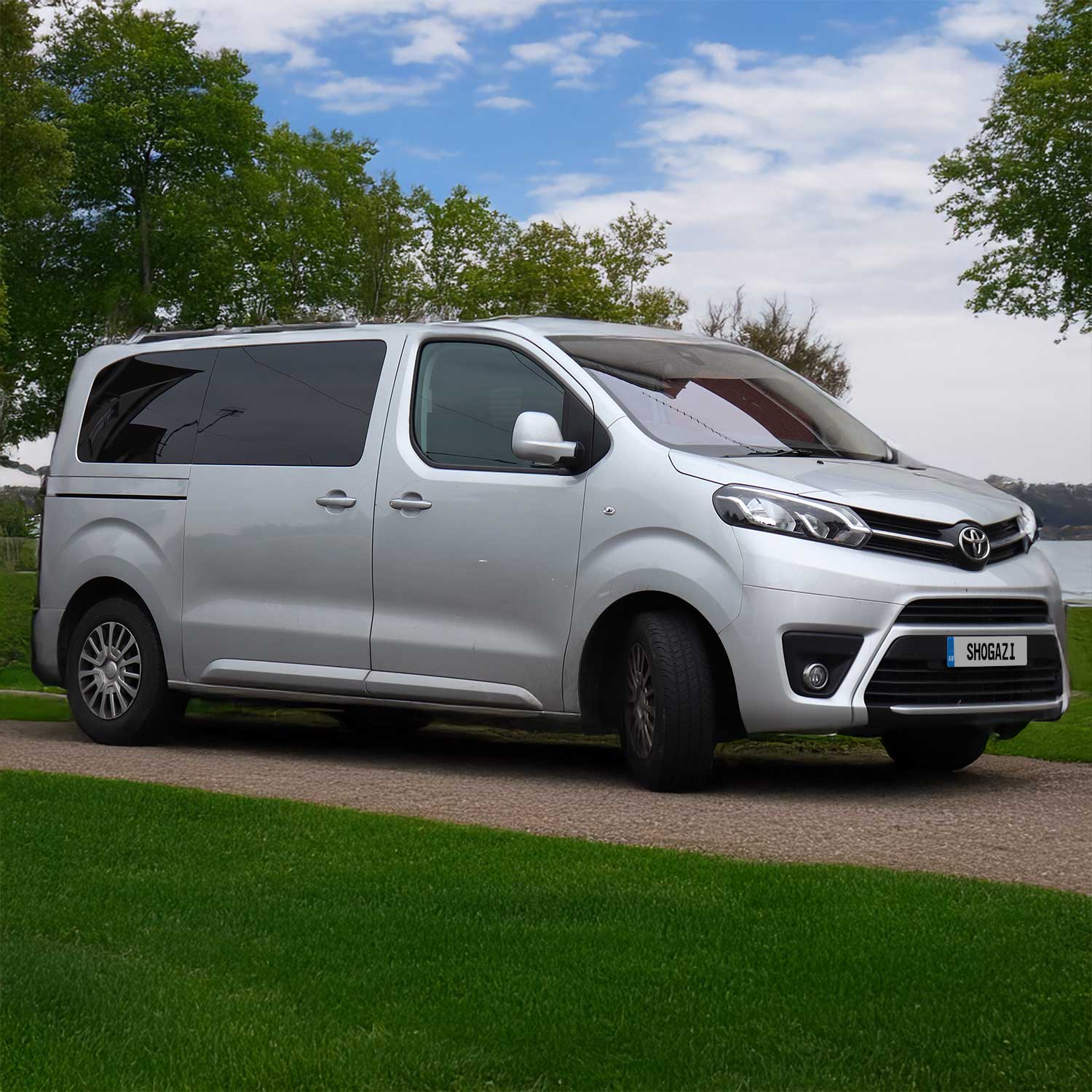 Toyota Proace Verso steht an einem See mit grüner Wiese im Vordergrund, blauem Himmel und weißen Wolken – Matratze 116x192 cm für komfortables Camping